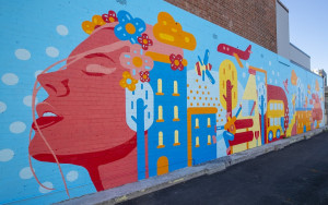 A cartoon-style mural painted using bright blues, oranges and reds, depicts a peaceful womans face and a range of buildings, transport and nature behind her.
