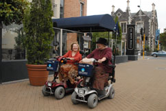 Two women on mobility scooters chat as they drive down a street